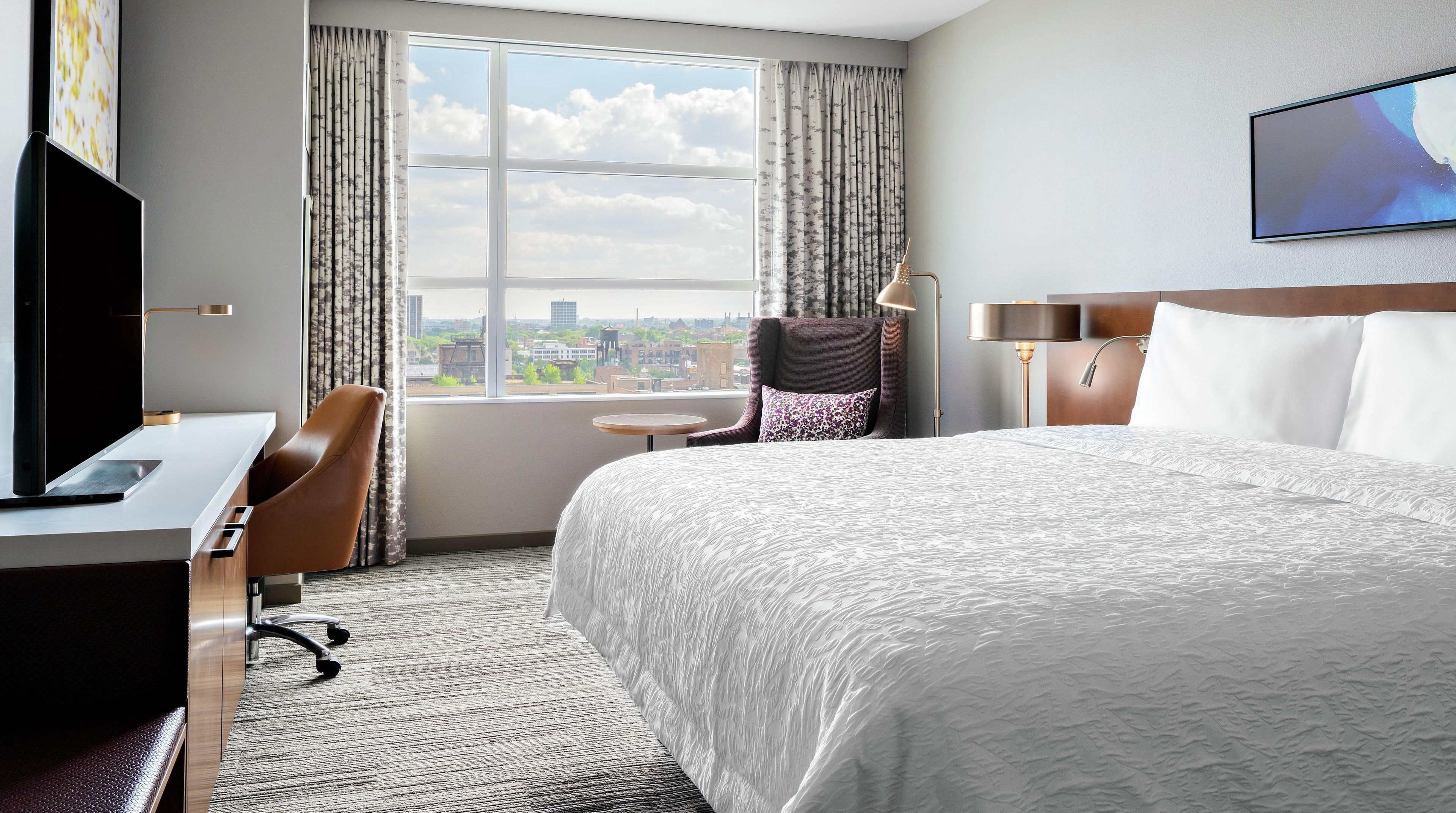 Quarto de hotel com uma cama grande, uma escrivaninha com computador, uma cadeira, uma mesinha e vista da cidade pela janela.