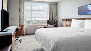 Quarto de hotel com uma cama grande, uma escrivaninha com computador, uma cadeira, uma mesinha e vista da cidade pela janela.