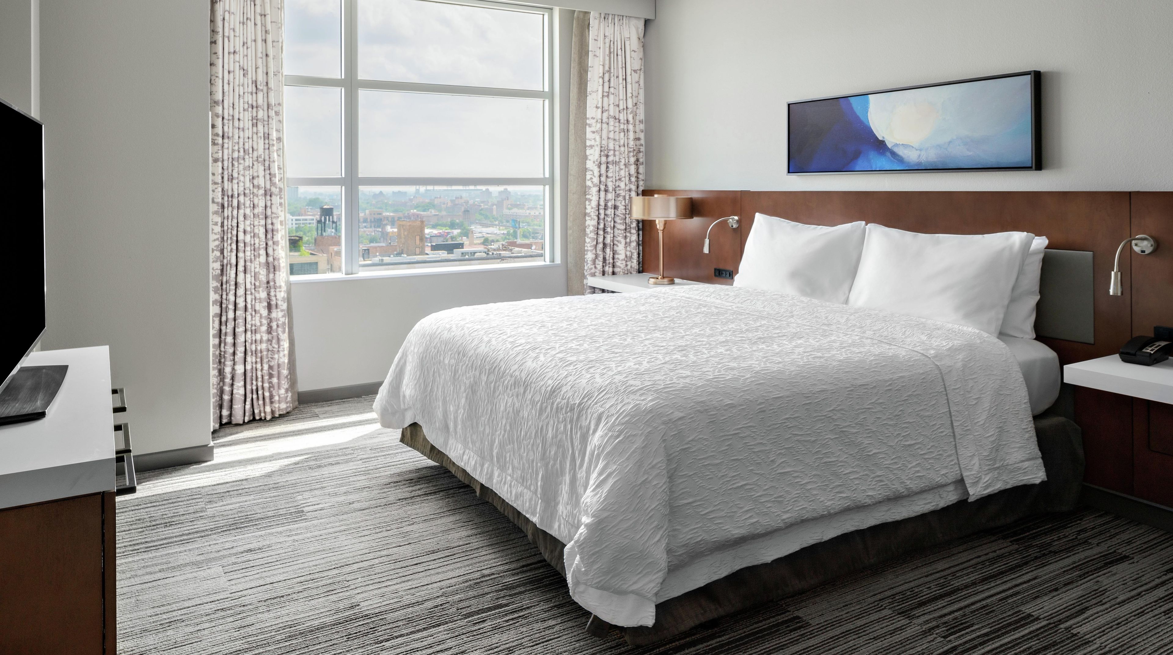A hotel room with a large bed, a desk with a computer, a window with a city view, and a painting on the wall.