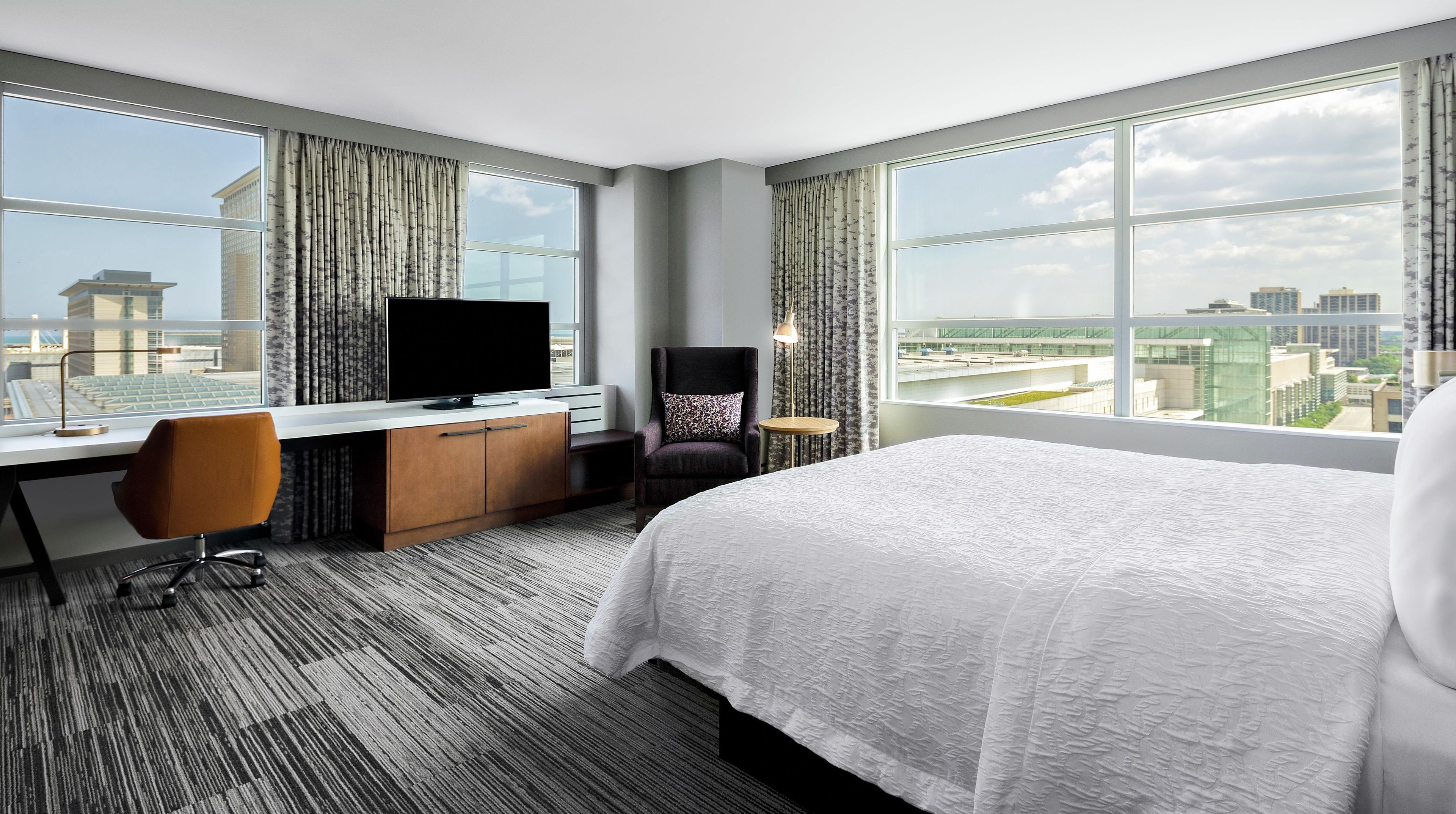 Habitación de hotel con una cama grande, un escritorio con televisor, una silla y vistas a la ciudad a través de grandes ventanales.