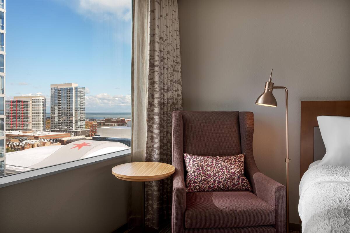 Une chambre d’hôtel avec vue sur la ville, une table ronde en bois, un fauteuil marron avec un coussin à motifs floraux et une lampe de chevet.