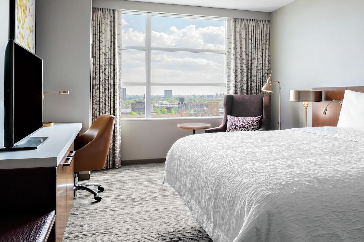 Une chambre d’hôtel avec un grand lit, un bureau avec un ordinateur, une chaise, une petite table et une vue sur la ville par la fenêtre.