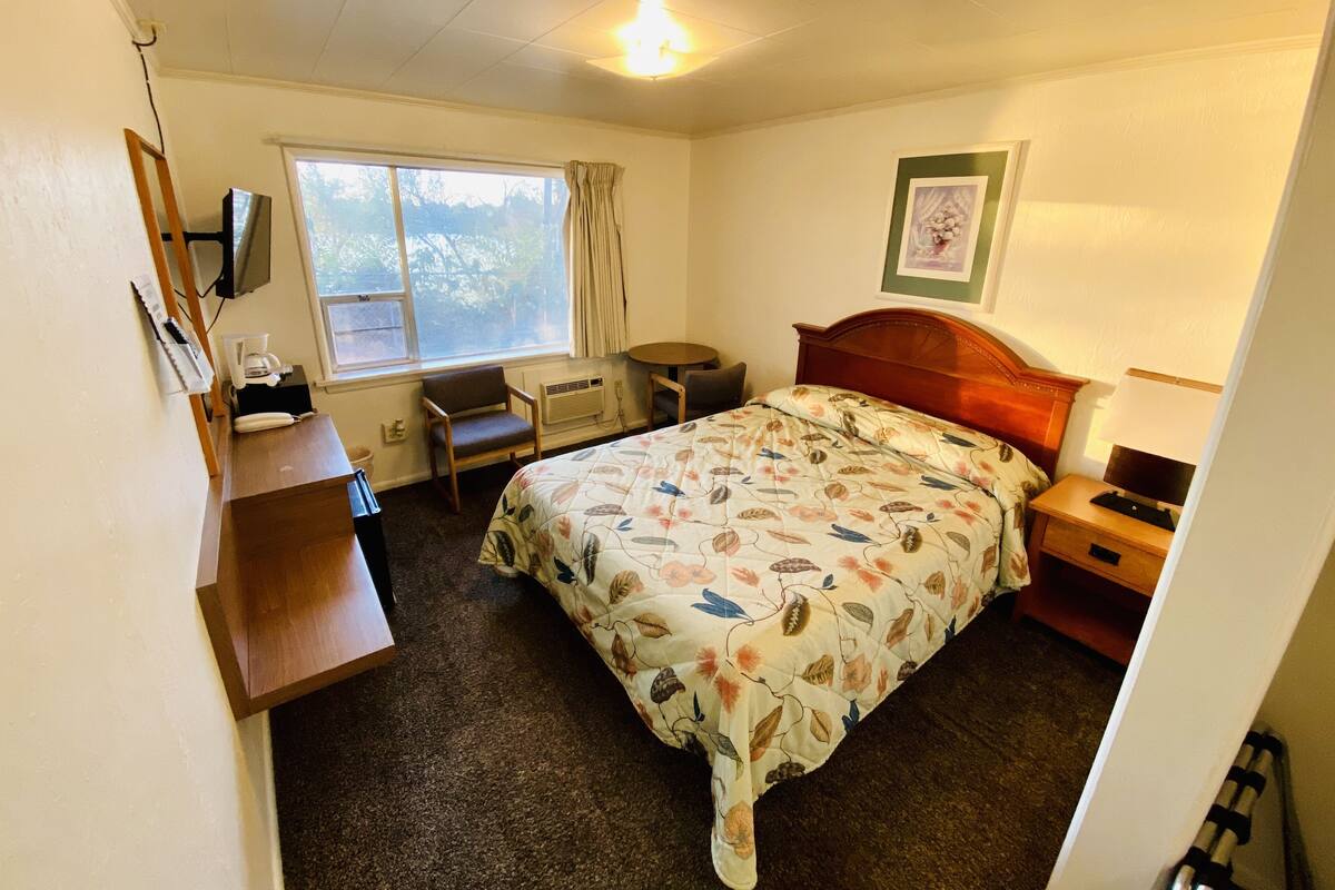 A hotel room with a bed, a desk, a chair, a window with curtains, and a framed picture on the wall.