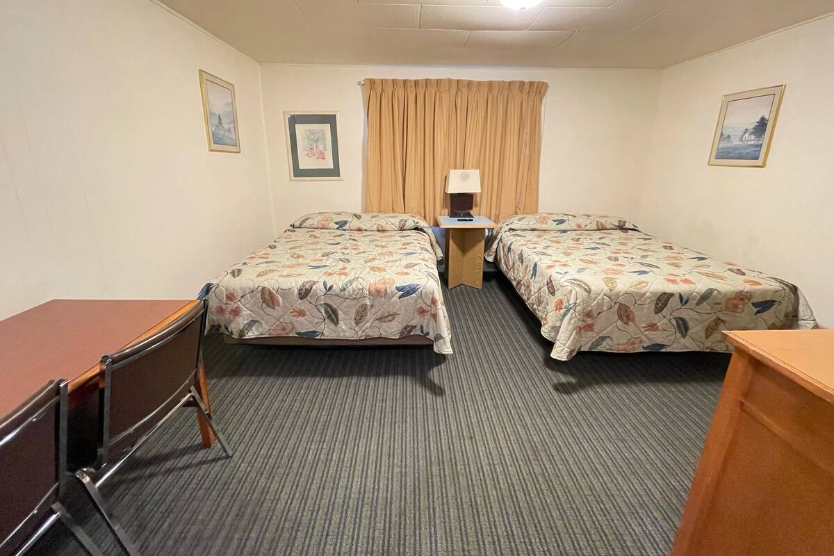 A hotel room with two single beds, a desk, a chair, a small table with a lamp, and a window with curtains.