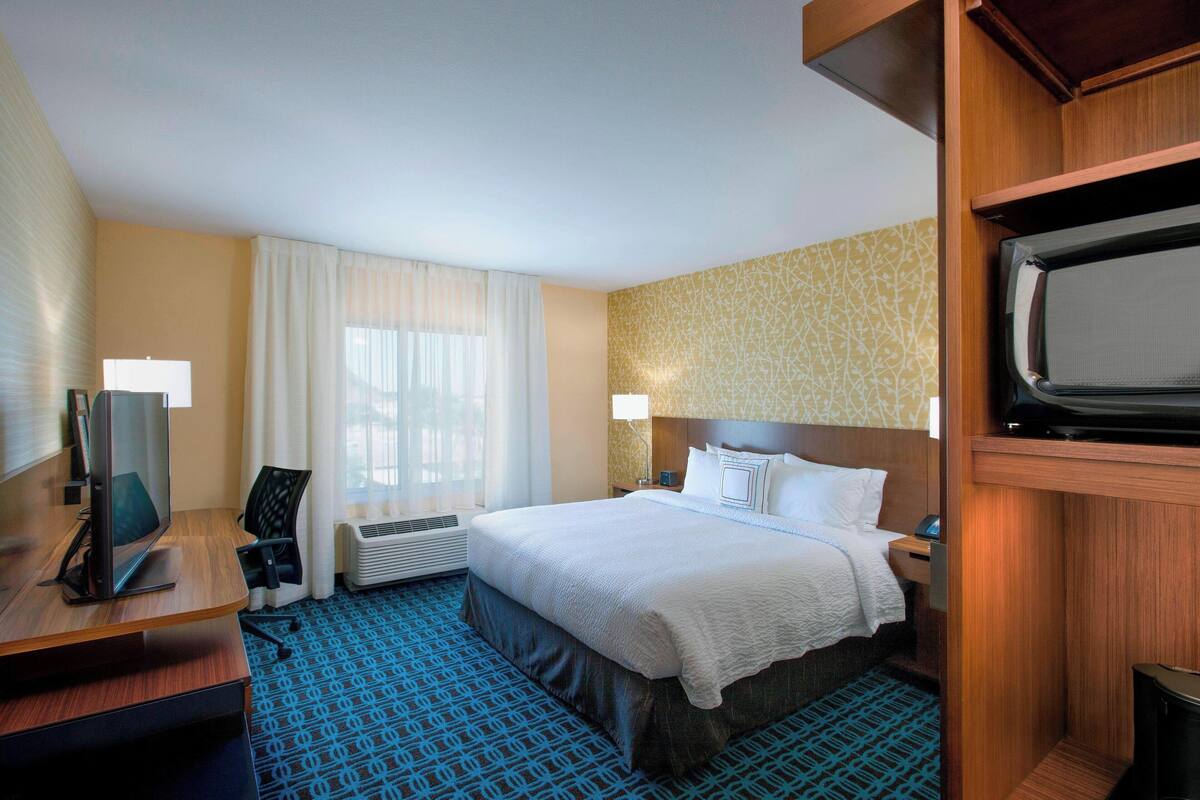 A hotel room with a bed, a desk with a computer, a TV, and a window with curtains.
