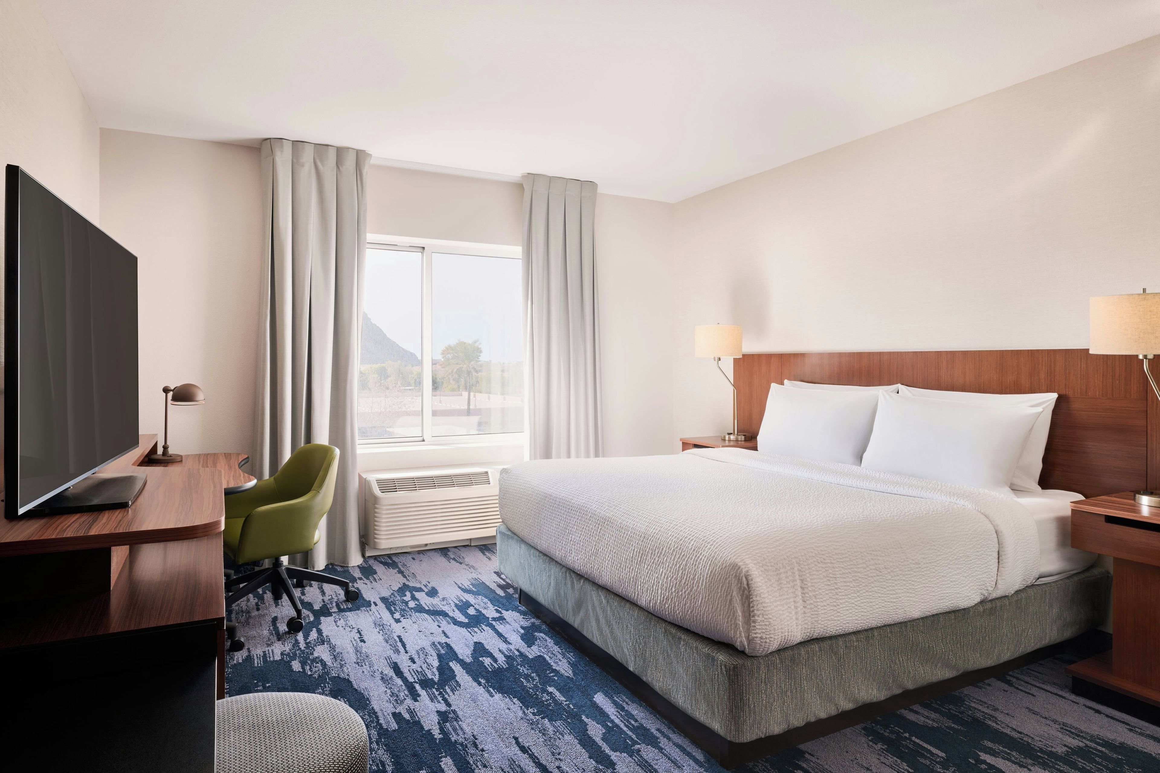 A hotel room with a bed, a desk with a flat-screen TV, a chair, a window with curtains, and a view of the outdoors.