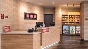 Un espace d’accueil d’hôtel avec un comptoir de réception en bois, une pancarte « Bonjour », une pancarte « Bienvenue » et une étagère proposant des collations pour les petites faims.