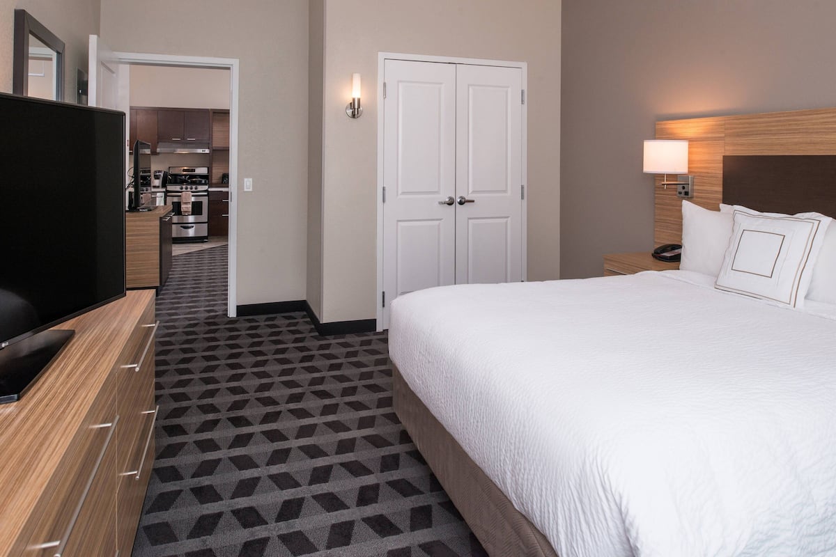 A hotel room with a large bed, a TV on a wooden dresser, a kitchen in the background, and a white door.