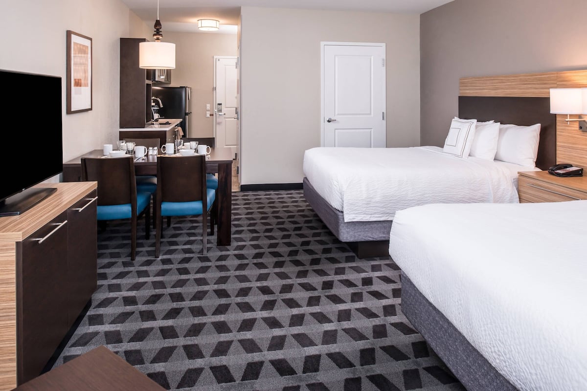 A hotel room with two beds, a desk with chairs, a TV, and a kitchen area.