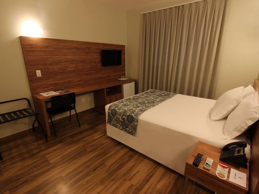 A hotel room with a bed, bedside table with a phone, a desk with a chair, a TV on the wall, and a window with curtains.