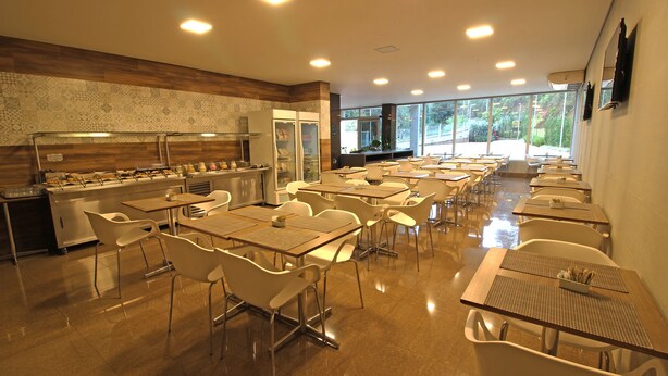 A spacious dining area with multiple tables and chairs, a buffet line, and large windows offering a view of the outdoors. - Ímpar Suítes Barão de Cocais (Barao de Cacais)