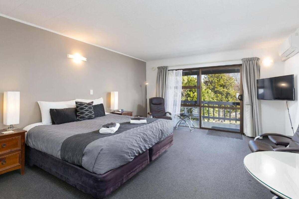 Habitación de hotel con una cama grande, un escritorio, una silla, televisión y balcón con vistas a árboles.