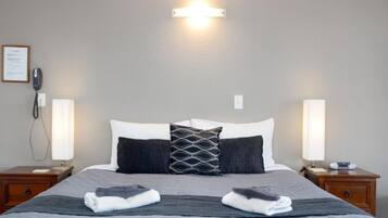 A neatly made double bed with a grey bedspread and two folded towels, flanked by two bedside tables with lamps, a framed fire safety notice, and a wall-mounted light fixture.