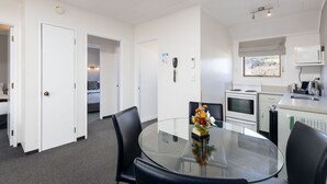 A hotel room with a glass dining table, black chairs, a flower vase, a kitchenette, and a bed in the background. - Kings Gate Motel (Nelson)