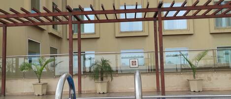 Una piscina con una pergola, piante in vaso e un edificio con ampie finestre sullo sfondo.