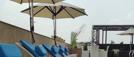 A row of lounge chairs with blue cushions and beige umbrellas, arranged on a rooftop terrace.