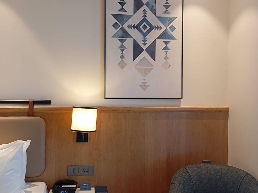 Una habitación de hotel con una cama, una mesita de noche con lámpara, una silla y una obra de arte colgada en la pared.