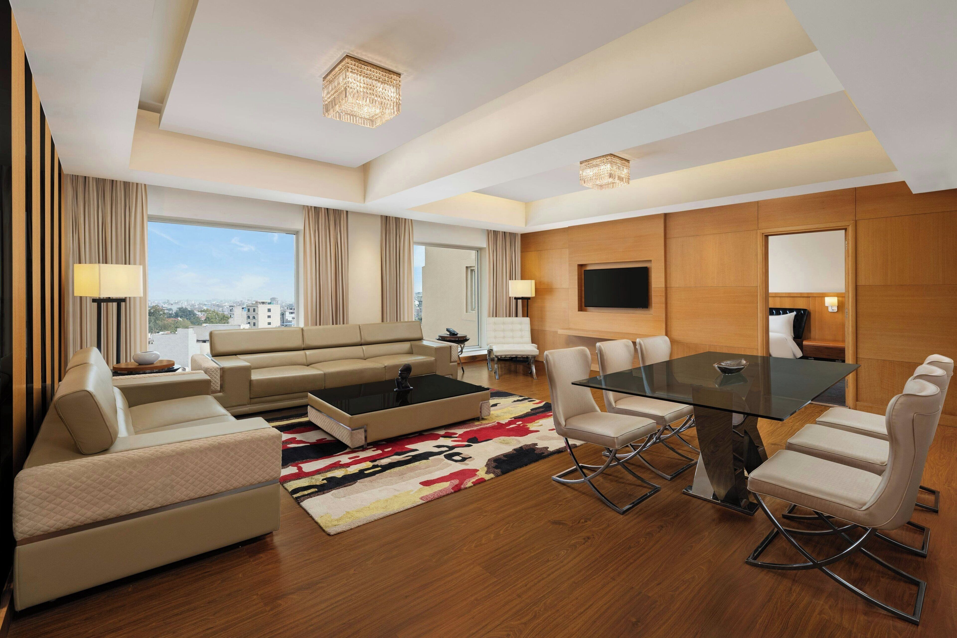 A modern hotel room with a sofa, coffee table, dining table, and chairs. There is a TV mounted on the wall and a large window offering a city view.