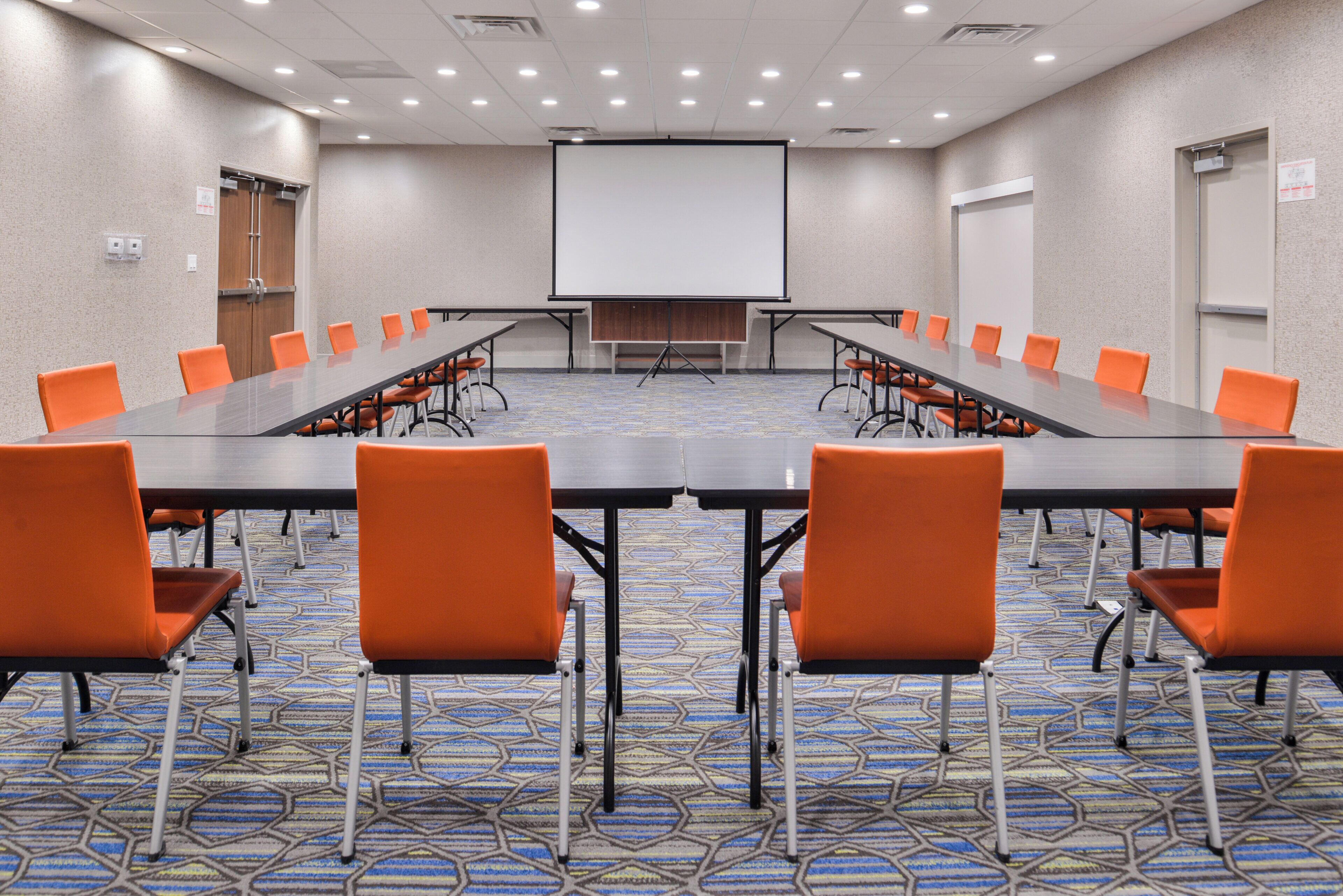 Una sala de conferencias con una larga mesa, sillas color naranja, una pantalla de proyector y piso alfombrado.