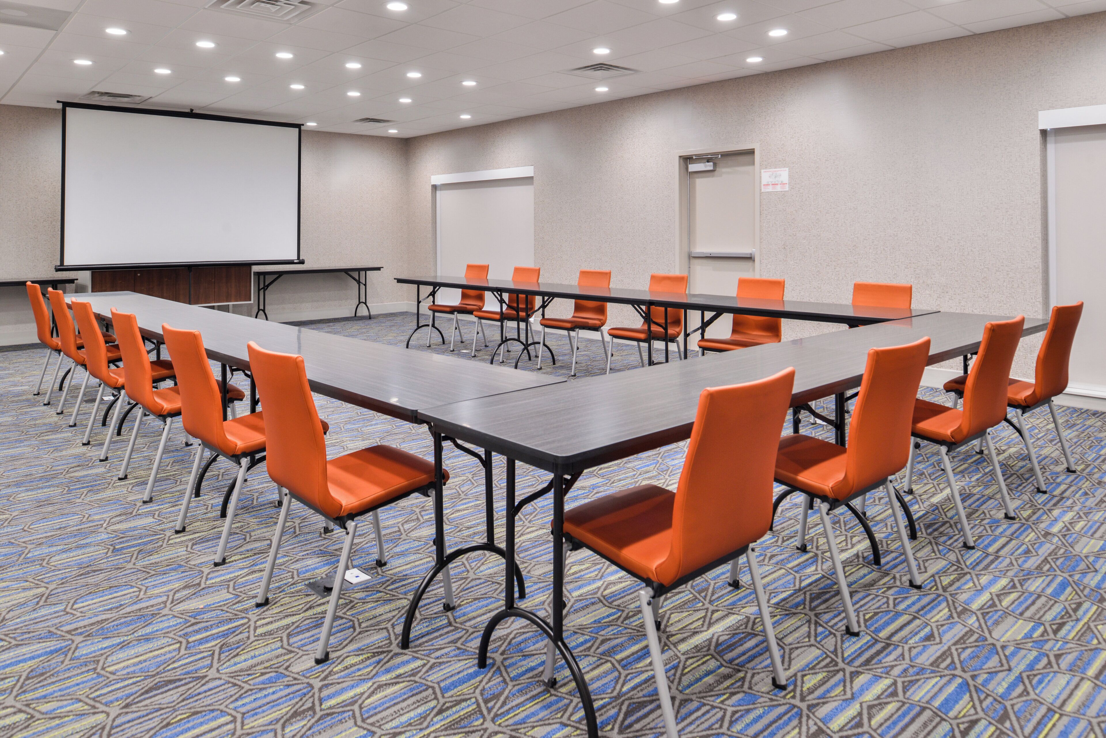 A conference room with a long table, orange chairs, a projector screen, and a carpeted floor.