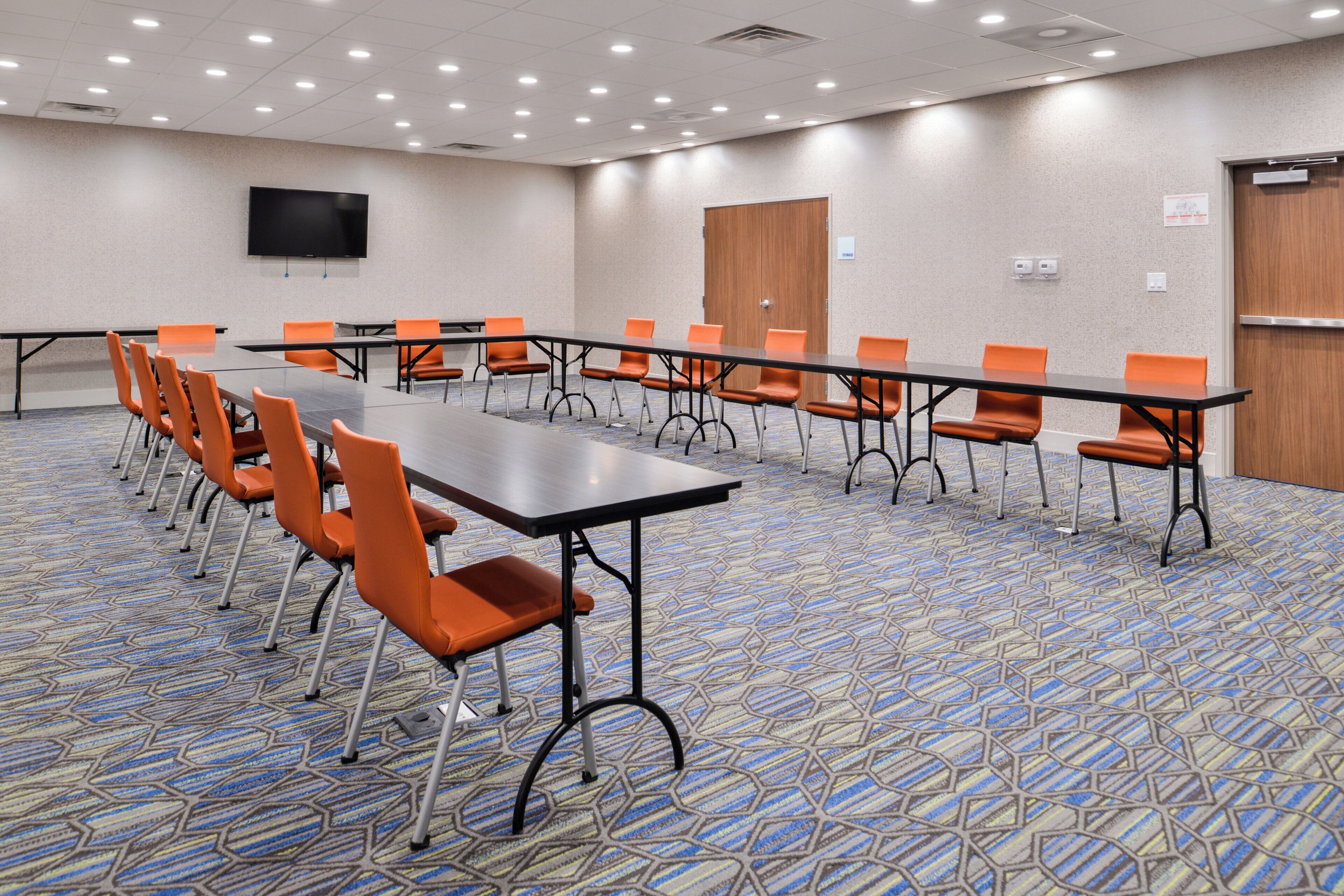 A conference room with a long table, orange chairs, a flat-screen TV on the wall, and a patterned carpet.