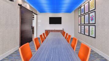 Un salón de conferencias con una mesa larga, sillas color naranja, un televisor de pantalla plana y decoración mural estampada.