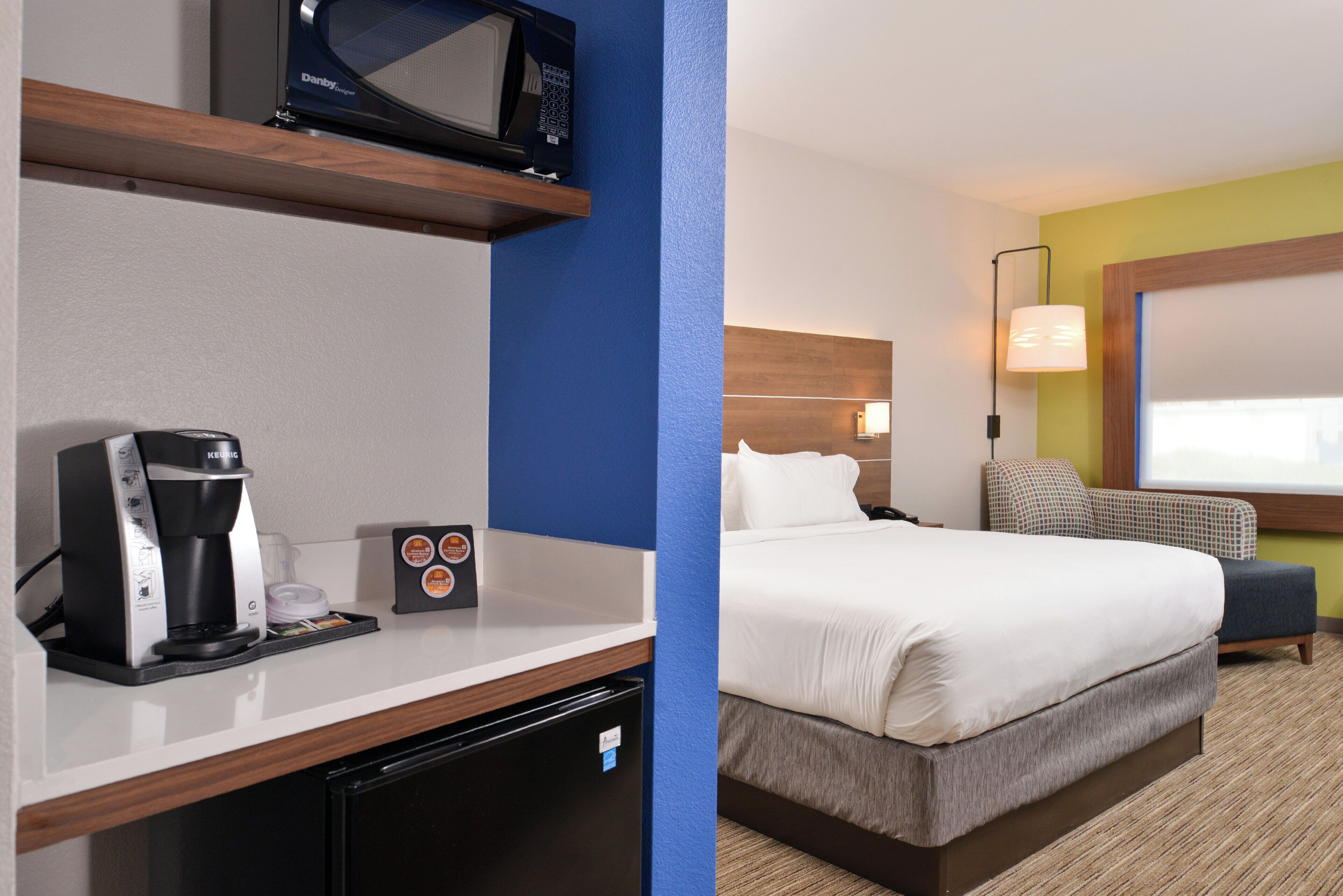 A hotel room with a bed, bedside table, coffee maker, microwave, and a lamp.