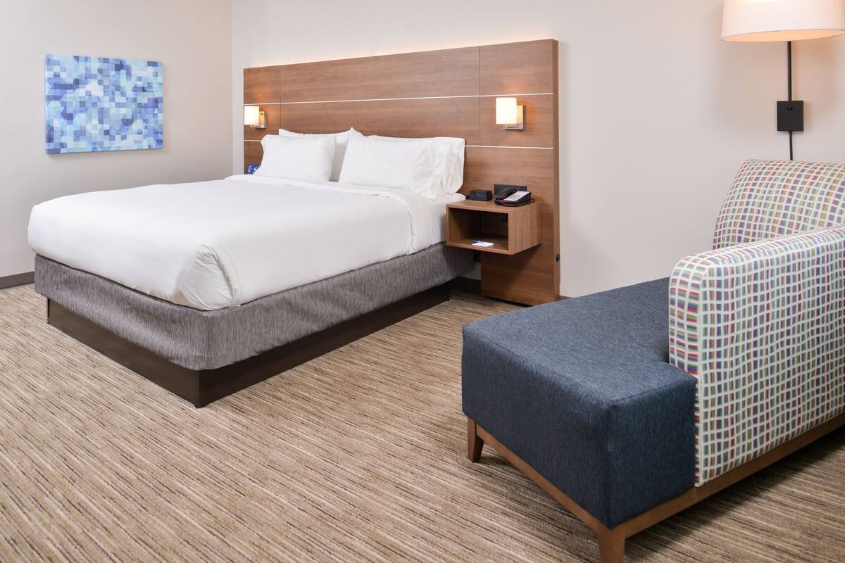 Una habitación de hotel con una cama grande, una mesita de noche con lámpara, un sillón estampado y una obra de arte colgada en la pared.