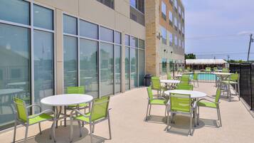 An outdoor patio area with round tables and chairs, a pool, and a building with large windows and a stone facade.
