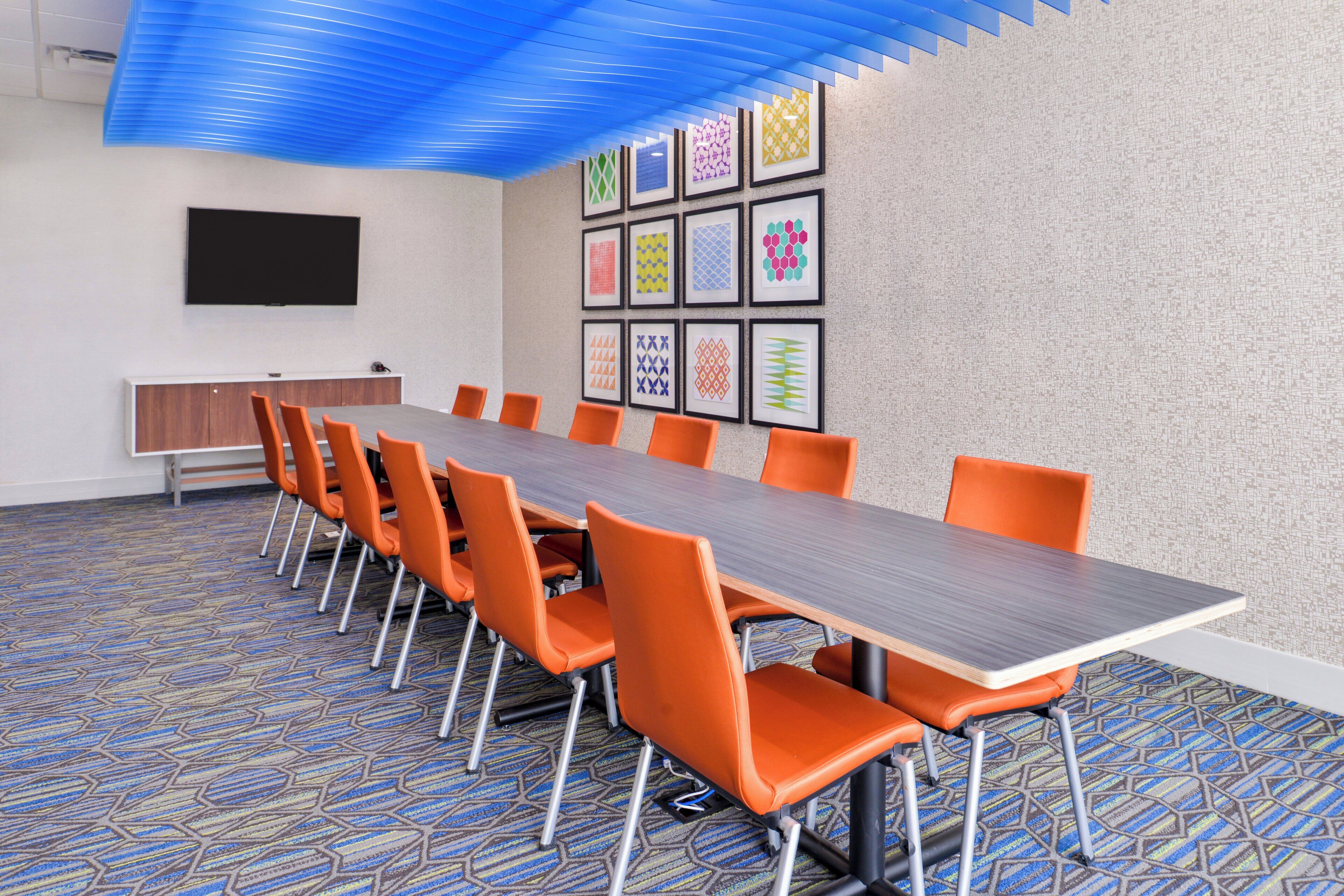 Une salle de conférence avec une longue table, des chaises orange, un téléviseur à écran plat et des œuvres d’art encadrées accrochées au mur.