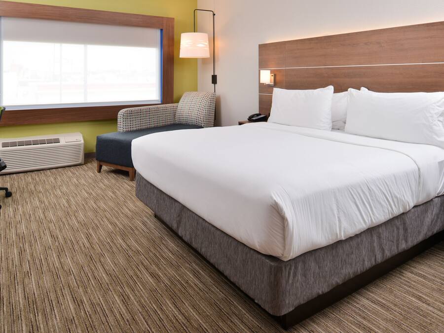Une chambre d’hôtel avec un grand lit, un bureau avec une chaise, une fenêtre avec des stores, une lampe et un appareil de climatisation.