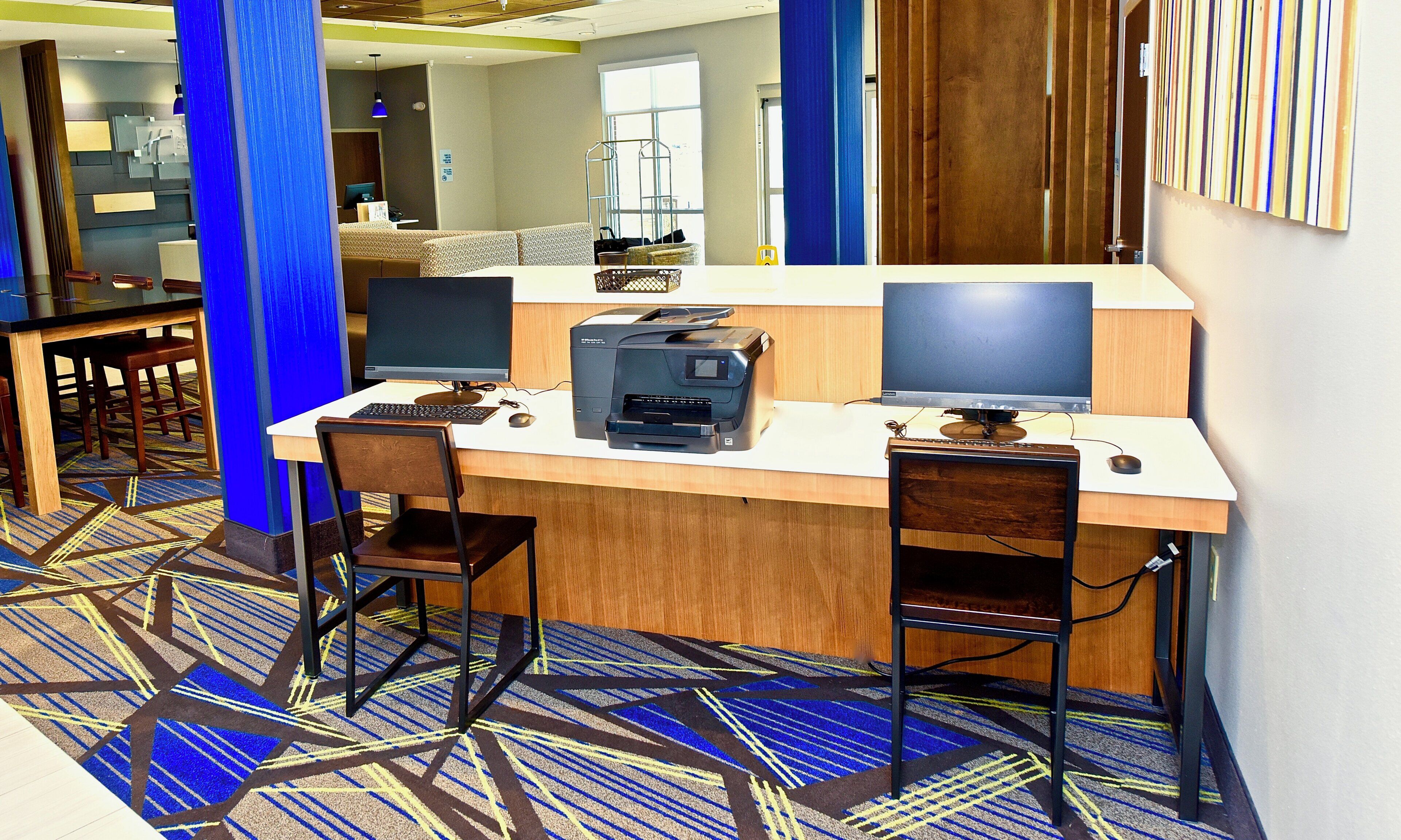 A workspace with two computer monitors, a printer, and a keyboard on a white desk. There are two chairs, a patterned carpet, and a blue pillar.