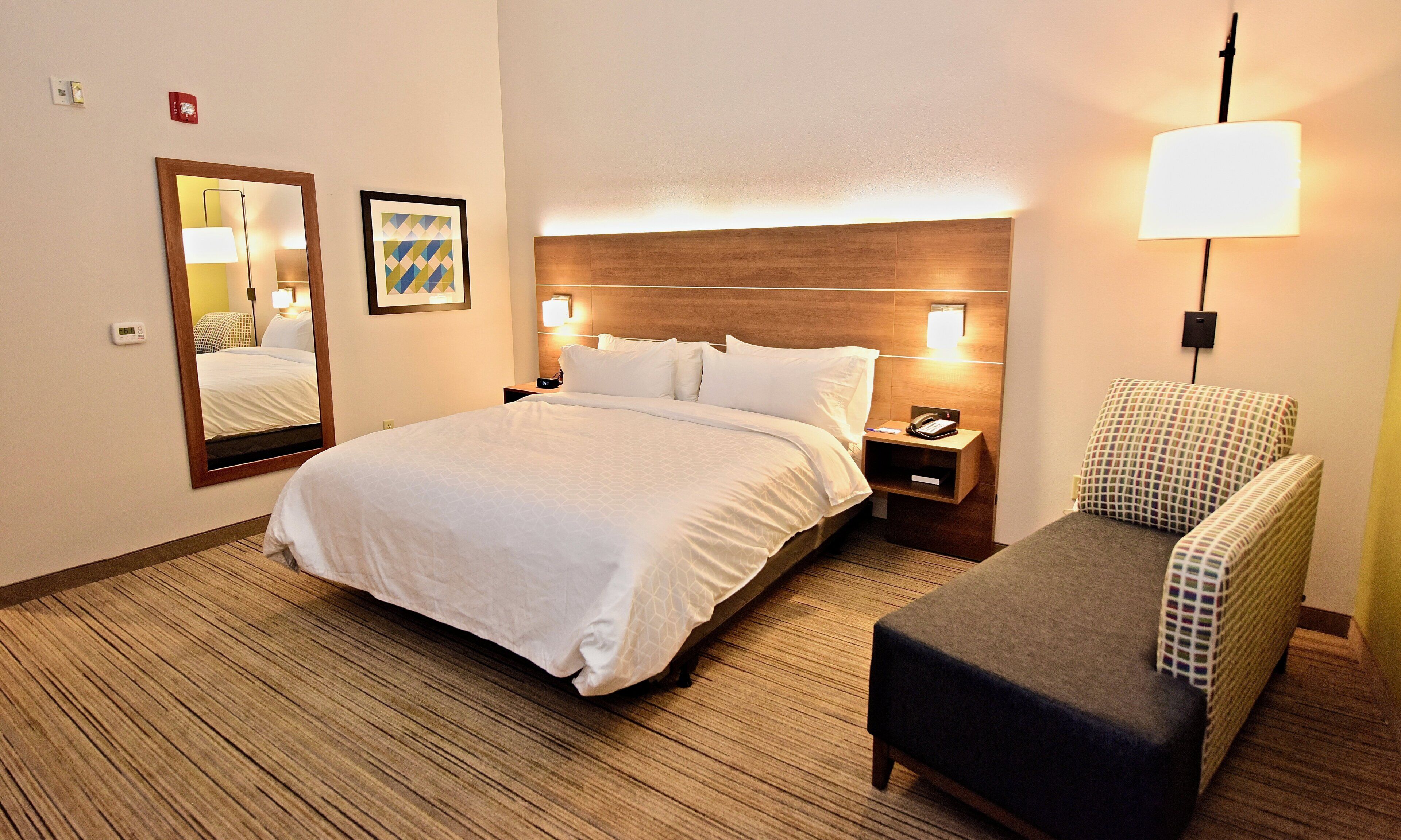 Una habitación de hotel con cama, mesita de noche, lámpara, silla y espejo.