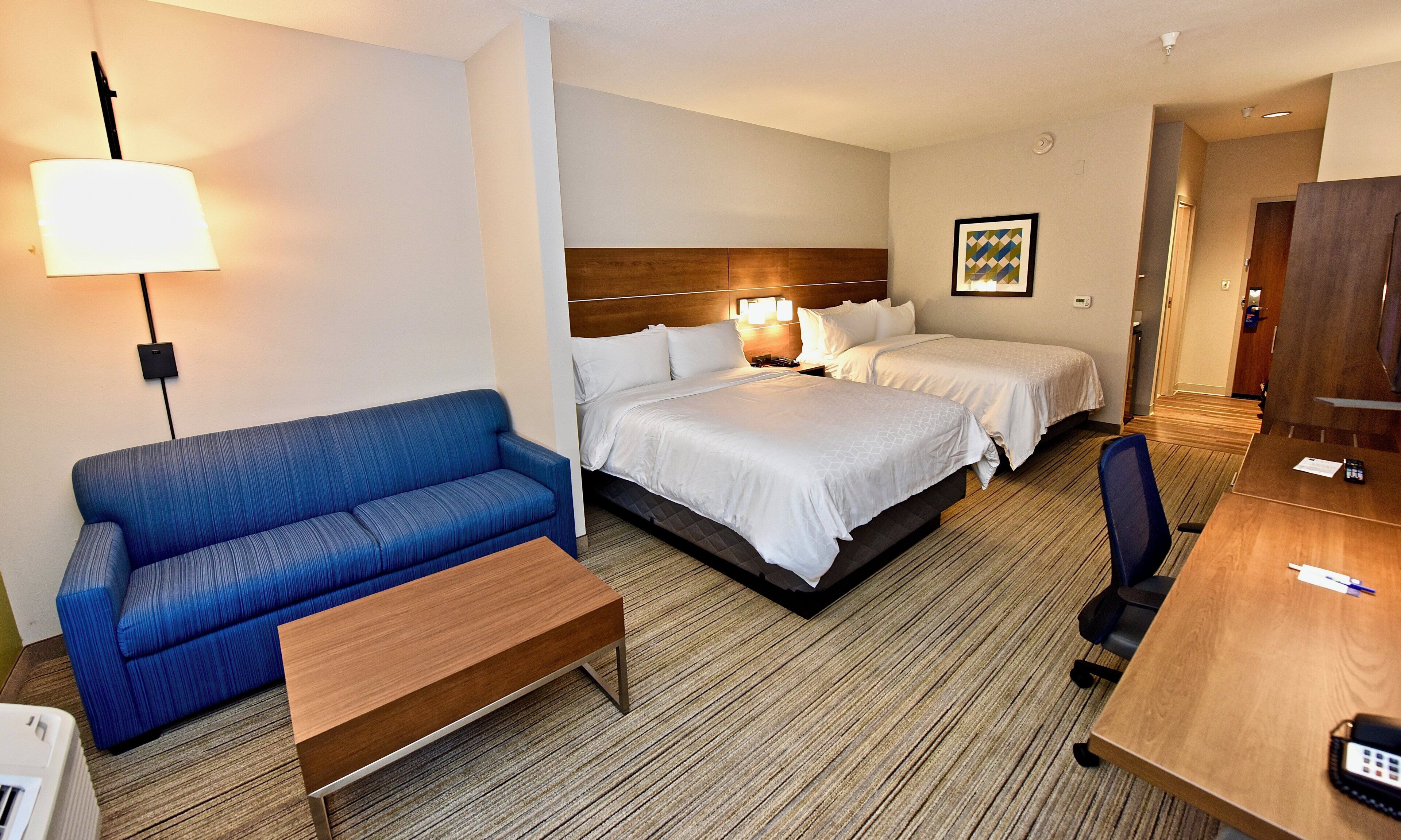 Een hotelkamer met een blauwe bank, een houten salontafel, twee bedden met witte lakens, een bureau met stoel en een wandlamp.
