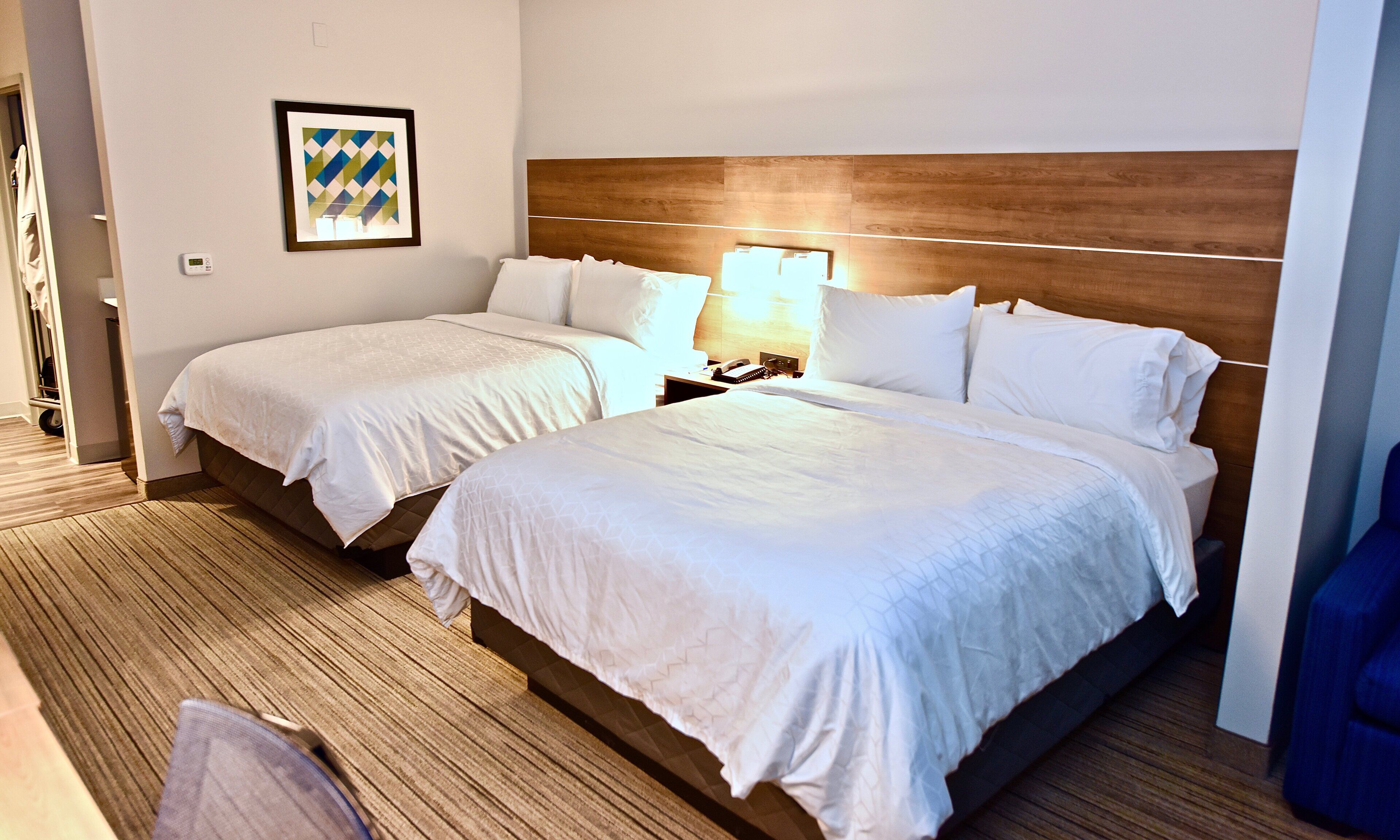 A hotel room with two beds, a wooden headboard, a bedside table with a lamp, and a framed picture on the wall.