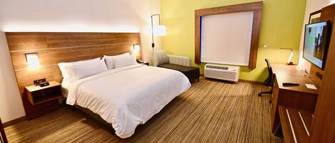 Una habitación de hotel con una cama grande, mesitas de noche, un escritorio con una silla, una lámpara de escritorio, un equipo de aire acondicionado y un televisor.