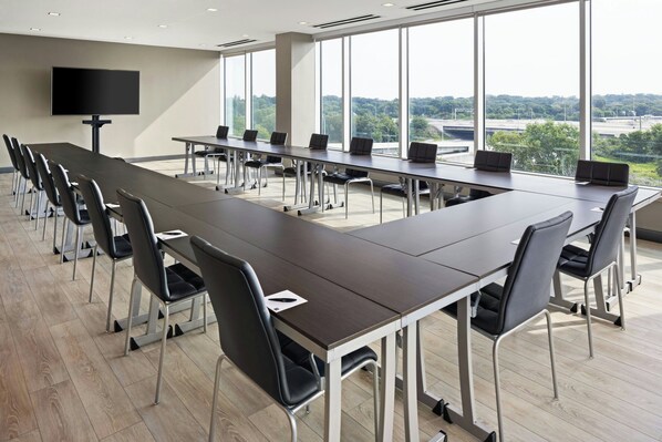 Un salón de conferencias con una larga mesa, sillas, un televisor de pantalla plana y ventanas amplias que ofrecen vistas a un puente y follaje.