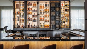 Un área de bar moderna con una variedad de botellas de licor y vasos, una isla central con asientos y una cocina elegante al fondo.