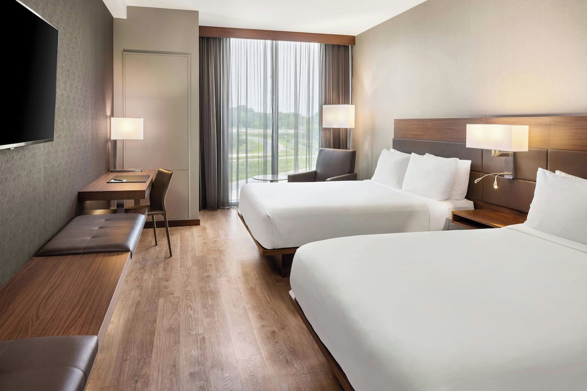 A hotel room with two beds, a desk, a chair, a TV, and a large window with curtains.