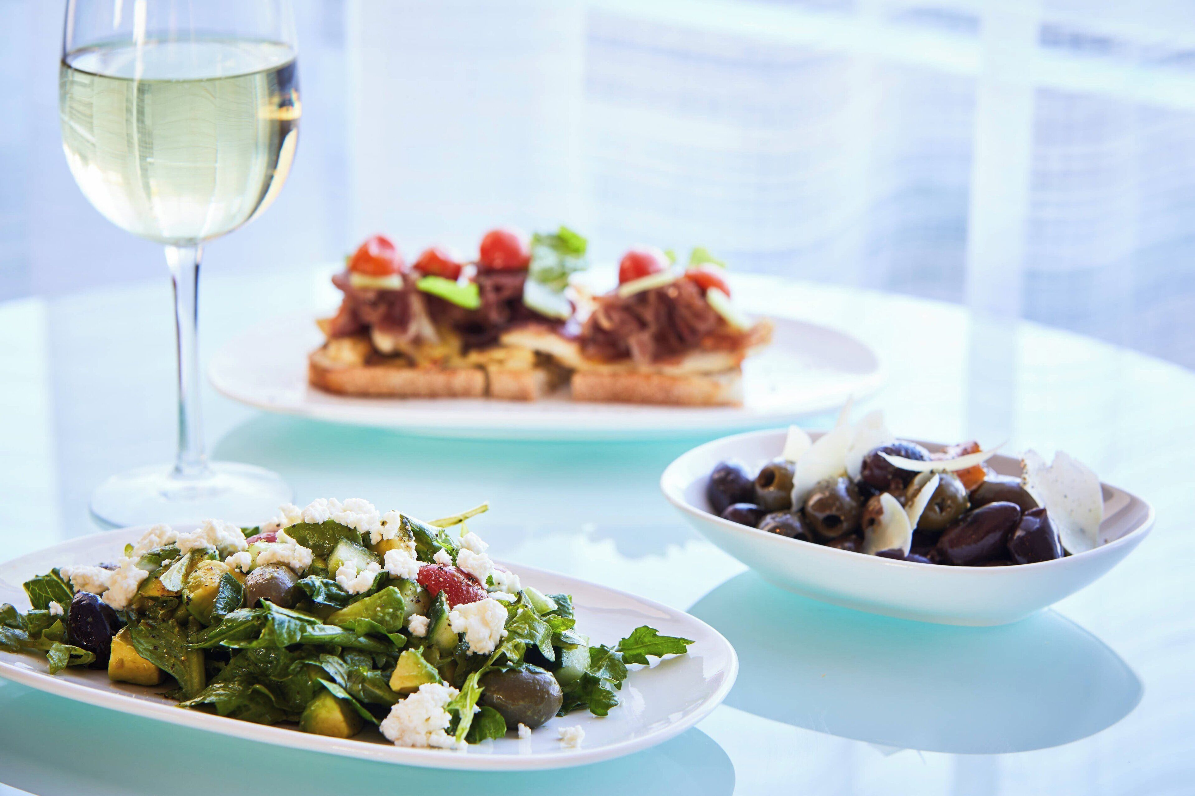 A table with a glass of white wine, a plate of salad, and a bowl of olives and feta cheese.