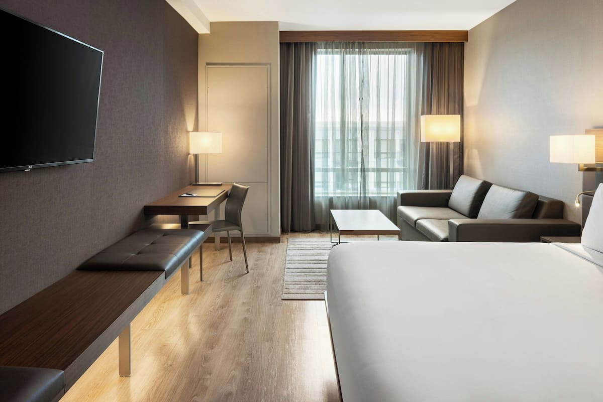 A modern hotel room with a large bed, a desk, a sofa, and a TV mounted on the wall.