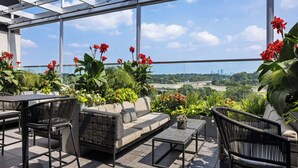 Un patio en la azotea con sofá, sillas y una mesa, rodeado de plantas y flores en macetas, y vistas de la ciudad a través de amplios ventanales.