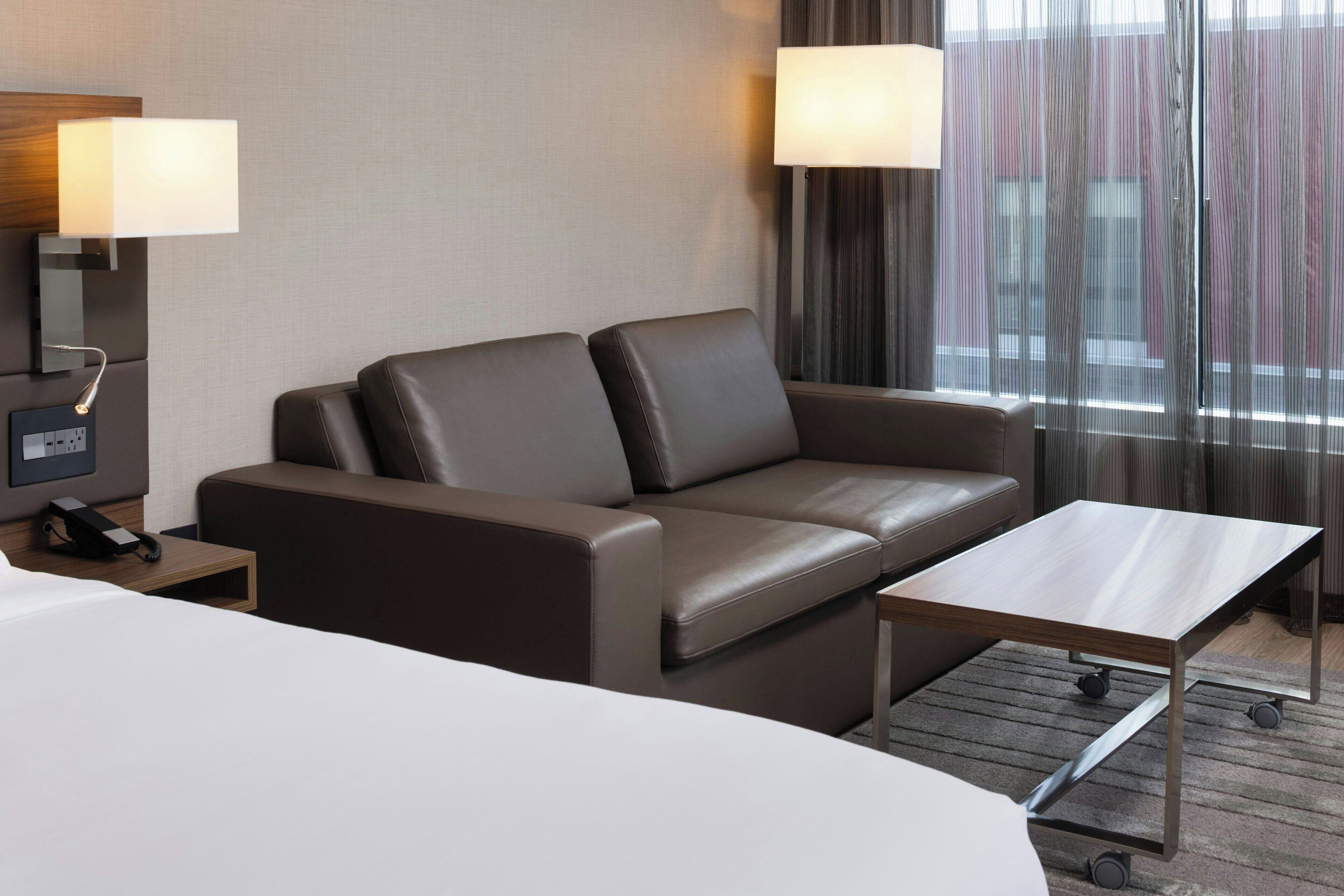 Een moderne hotelkamer met een leren bank, een houten salontafel, een nachtkastje met een lamp en een groot raam met gordijnen.