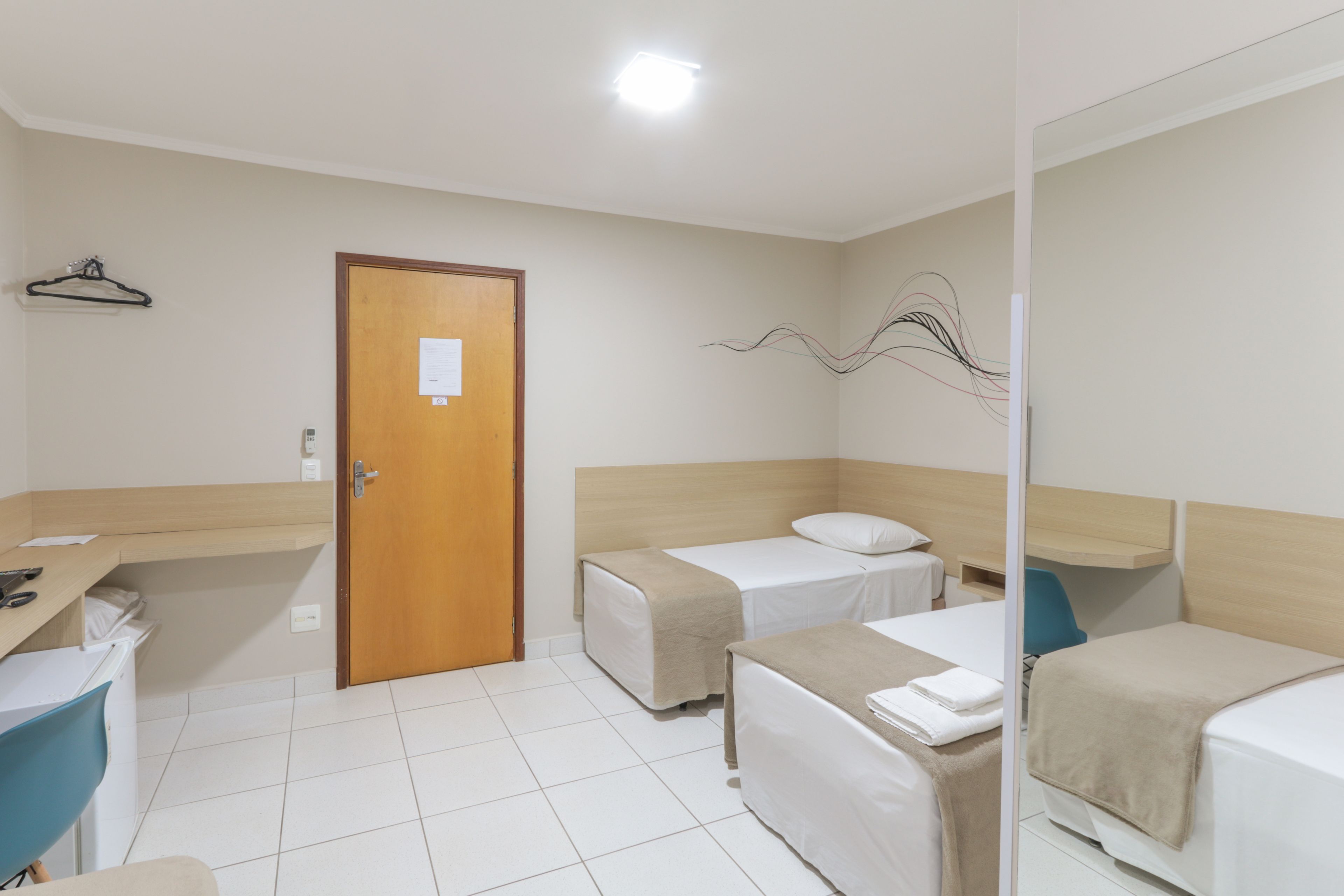 A hotel room with two single beds, a desk with a chair, a wooden door, and a wall-mounted art piece.