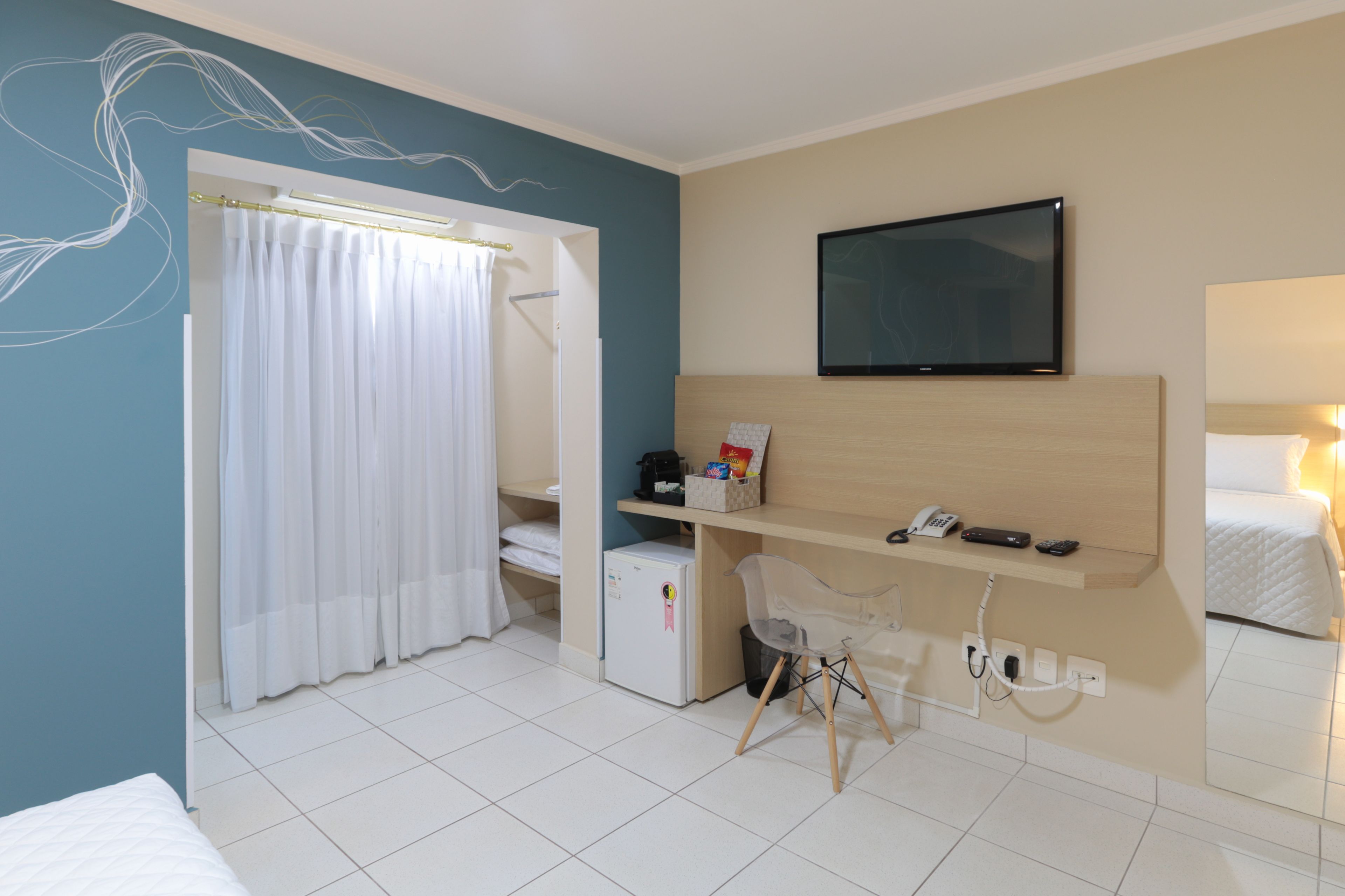 A hotel room with a bed, a desk with a chair, a TV, a mini-fridge, and a shower area.