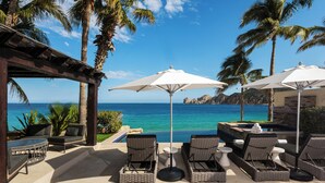 A poolside area with lounge chairs and umbrellas, overlooking a clear blue sea and palm trees.