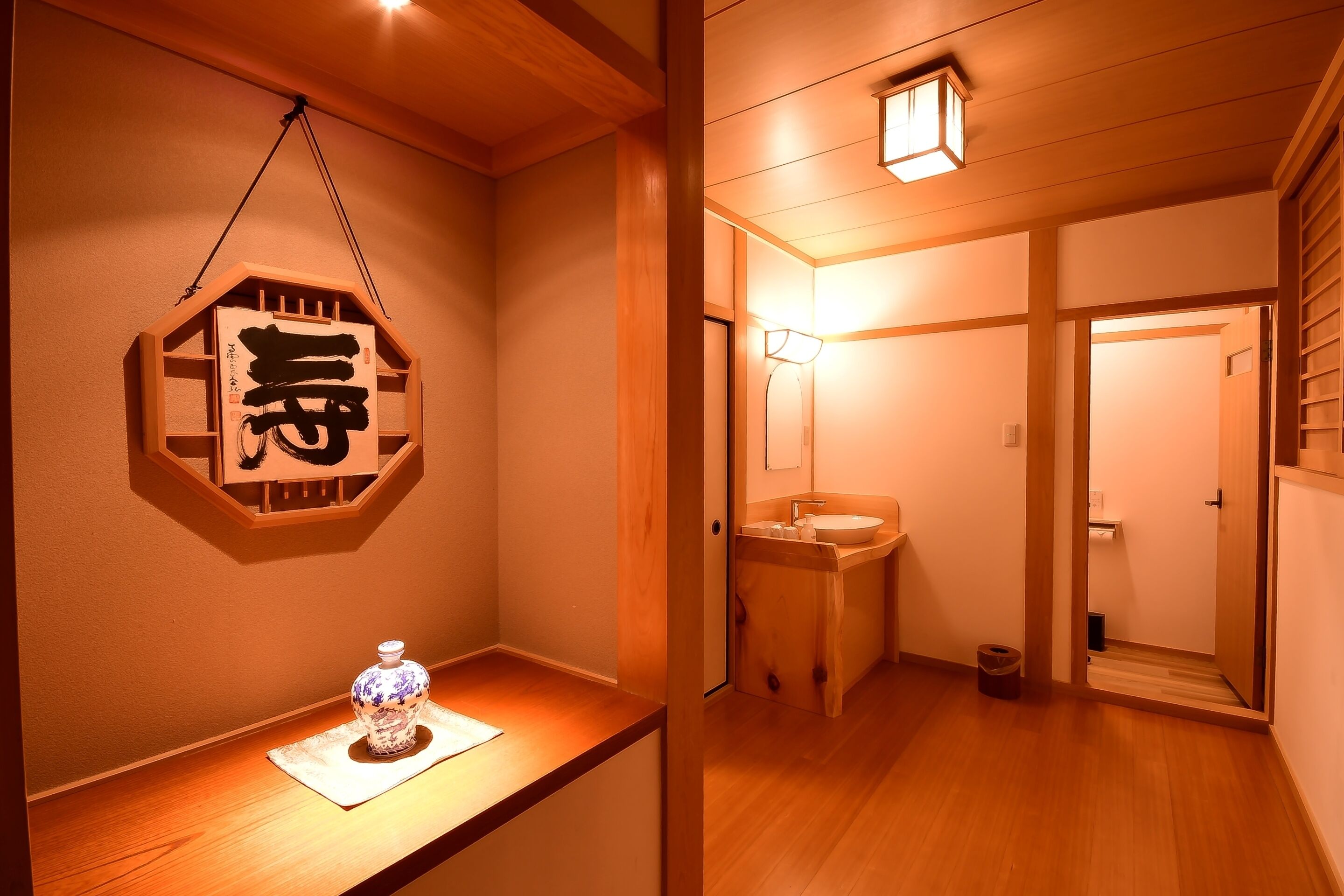 Una habitación japonesa tradicional con piso de madera, un estante de madera con un objeto decorativo, una obra de caligrafía colgada y una puerta de madera que da a otra habitación.