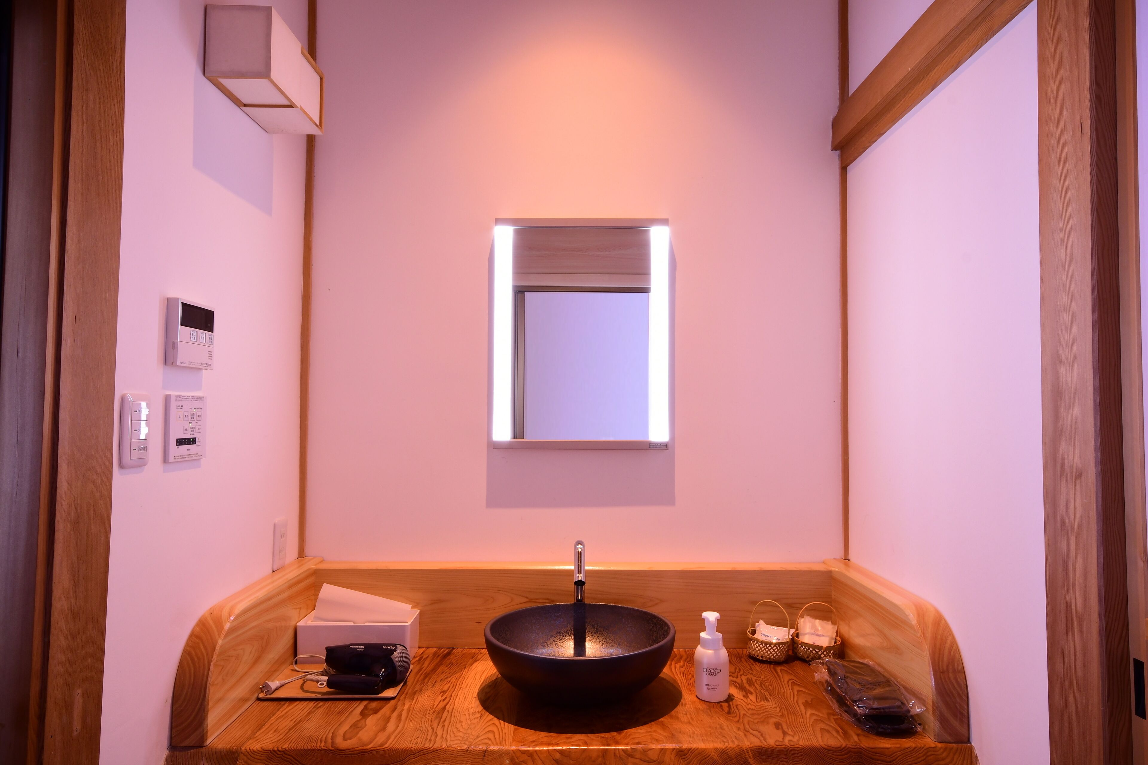 A modern bathroom with a wooden vanity, a black vessel sink, a small window, and a wall-mounted light fixture.