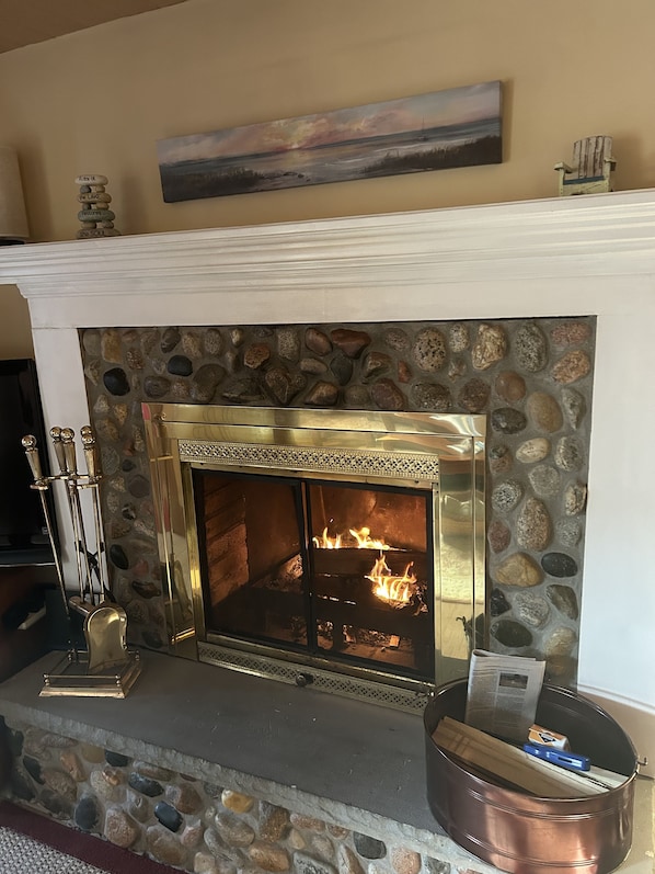 A fireplace with a stone surround and a metal grate, a mantelpiece with a painting, and a rustic chair. - Cozy relaxing getaway - Great trails, sunsets, dunes, jacuzzi tub, beach (Charlevoix)