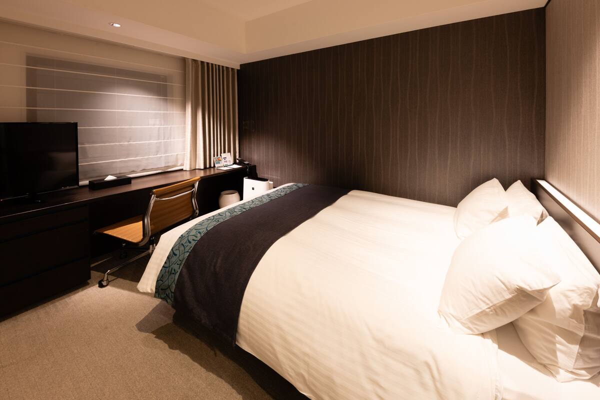 A hotel room with a large bed, a desk with a chair, a television, and a window with curtains.